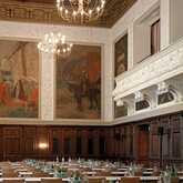 The elegant Huebner Saal features ornate chandeliers, historical murals, and neatly arranged tables with bottled beverages.