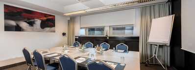 A modern meeting room with a large table, blue chairs, and a flip chart, featuring a vibrant abstract artwork on the wall.