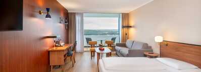A modern hotel room features a cozy bed, a sofa, and a desk, with a scenic balcony view.