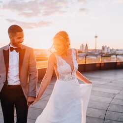 A couple in elegant attire joyfully holds hands on a rooftop with a stunning sunset city view.