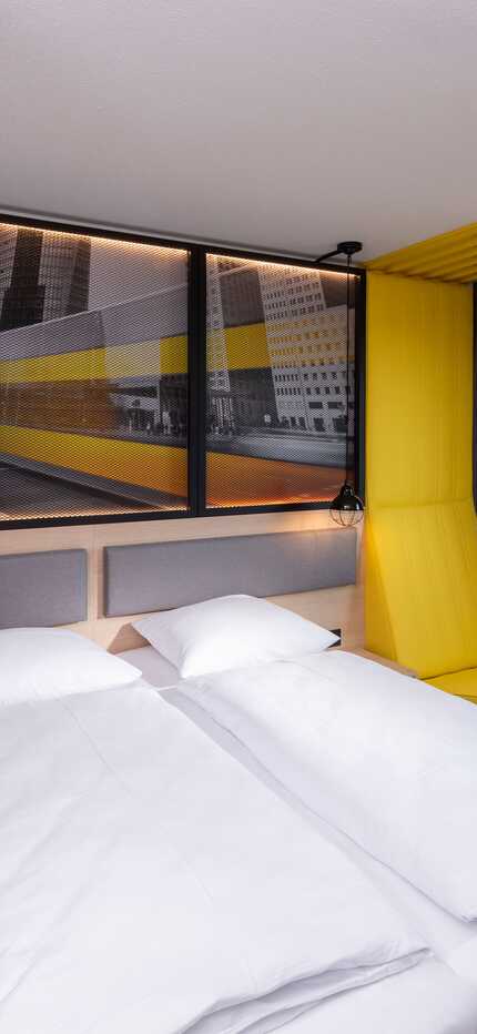 A modern hotel room features a large bed, vibrant yellow accents, and a window view of a riverside cityscape.