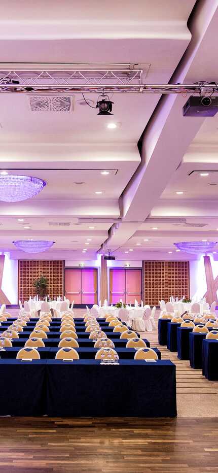A spacious conference room is set up with rows of tables and chairs, illuminated by purple lighting for a formal event.