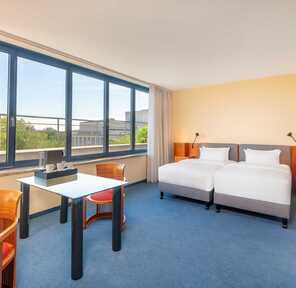 A bright hotel room features twin beds, a desk, and large windows offering a scenic view of trees and distant buildings.