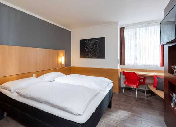 A modern standard room features a neatly made bed, stylish artwork, and a cozy seating area by the window.