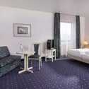 A spacious superior room features a cozy double bed, a patterned sofa, and a small table on a plush carpet.