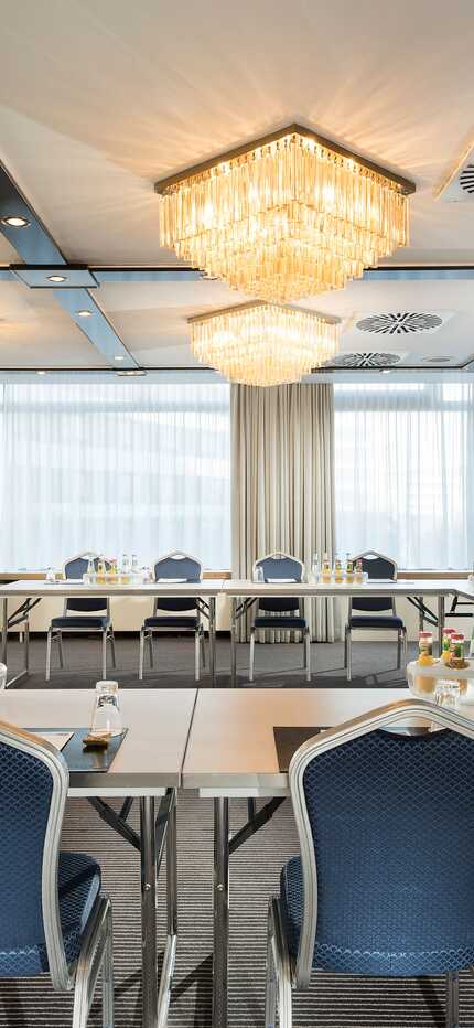 The meeting room features elegant chandeliers and a U-shaped table setup with chairs and refreshments.