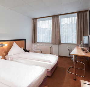 Ein modernes Doppelzimmer mit zwei Einzelbetten, einem Schreibtisch und großen Fenstern mit Vorhängen.