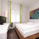 Ein modernes, helles Hotelzimmer mit Doppelbett, Flachbildfernseher und farbenfrohem Wandbild lädt zum Entspannen ein.