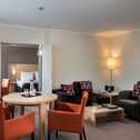 A modern suite features a cozy living area with vibrant orange chairs and a view into a neatly arranged bedroom.