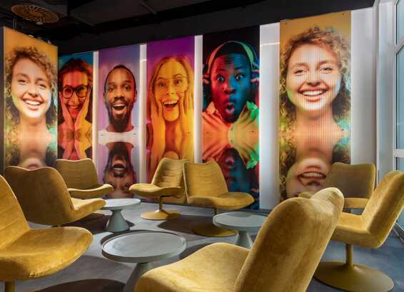 A modern lobby features vibrant floor-to-ceiling portraits and stylish yellow chairs arranged around round tables.