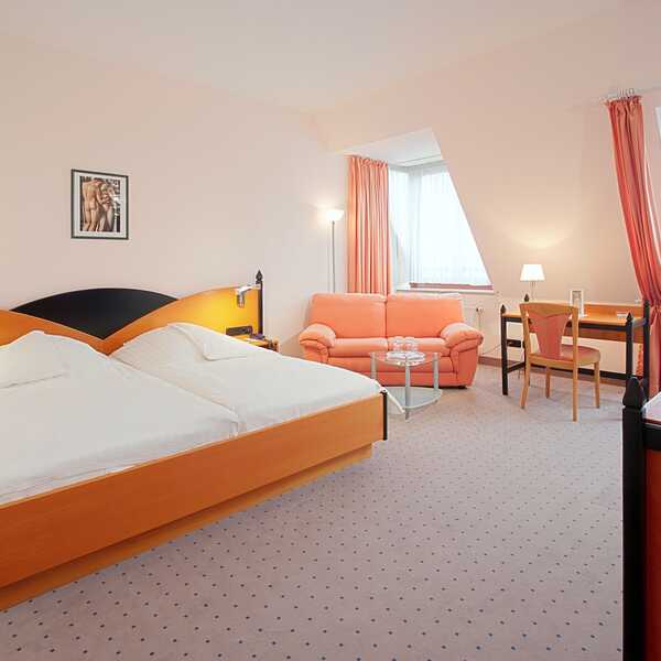 Ein gemütliches Doppelzimmer mit zwei Betten, einer rosa Couch und einem Schreibtisch mit Fernseher in einem hellen Raum.