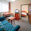 A cozy hotel suite features a blue patterned sofa, a wooden table with glasses, and a view into a bedroom with a neatly made bed.