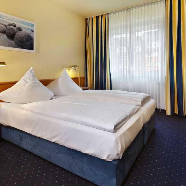 Gemütliches Doppelzimmer mit blau-gelben Vorhängen, einem großen Bett und einem Bild von Steinen an der Wand.