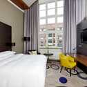 A modern hotel room features a cozy bed, a vibrant yellow chair, and a large window overlooking an urban view.