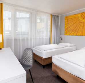 A modern hotel room features two neatly made twin beds, a vibrant yellow artwork, and a desk with a TV and lamp.