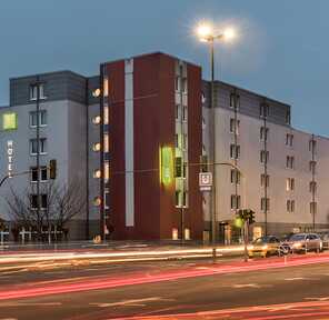 Cars drive past a modern hotel building at dusk, with bright streetlights illuminating the exterior.