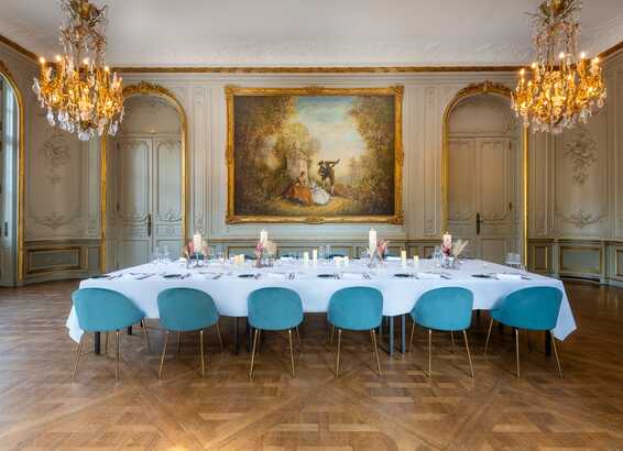 Ein eleganter Speisesaal mit einem langen Tisch, blauen Stühlen, opulenten Kronleuchtern und einem großen Gemälde an der Wand.