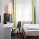 Ein helles Hotelzimmer mit einem Einzelbett, einem Schreibtisch und einem Fernseher neben einem großen Fenster.