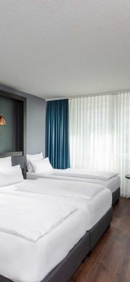 A modern hotel room features twin beds, stylish lighting, and a cozy seating area near large windows with blue curtains.