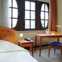 A cozy single room features a bed, a wooden desk, and an arched window overlooking the cityscape.