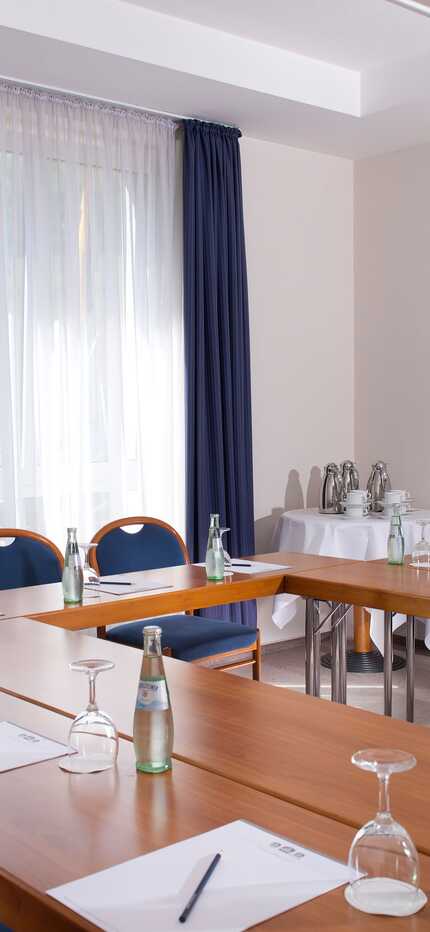 A well-lit meeting room with arranged tables, chairs, notepads, and bottled water awaits attendees.