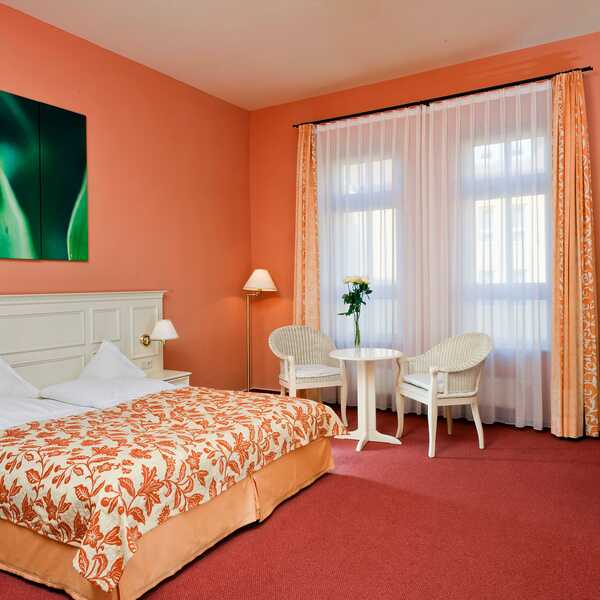 Gemütliches Zimmer mit einem großen Bett, einem kleinen Tisch mit zwei Stühlen und einem beruhigenden grünen Kunstwerk an der Wand.