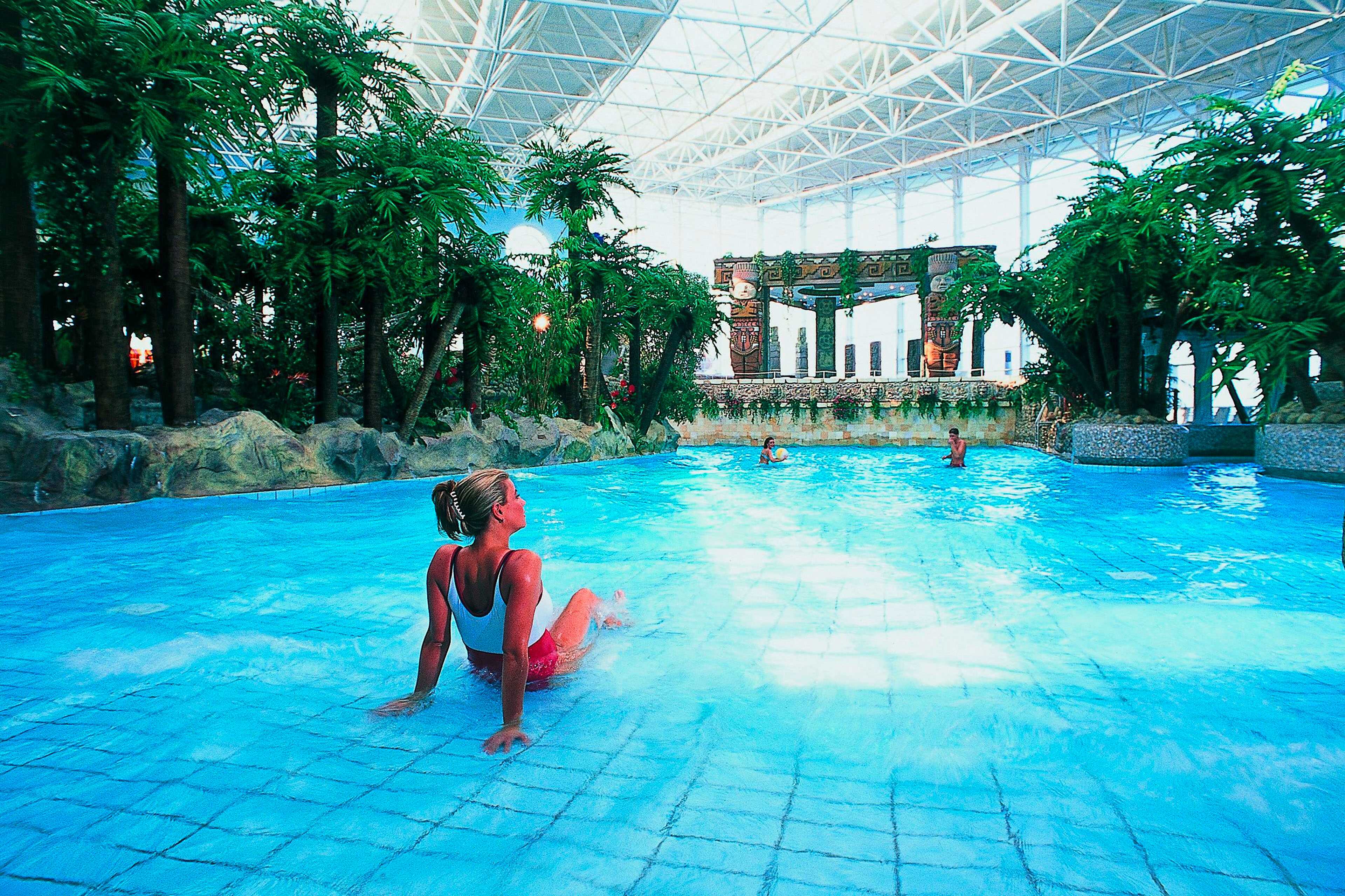 Eine Frau entspannt sich am Rand eines großen, tropischen Indoor-Pools, umgeben von Palmen und exotischer Dekoration.