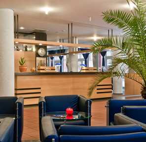 The hotel lobby features modern blue seating, a large potted palm, and a welcoming reception desk.