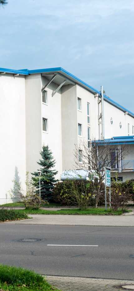 A modern residential building with blue accents stands surrounded by greenery on a quiet street.
