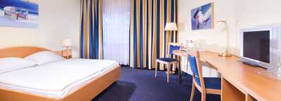 A cozy room features a comfortable bed, a desk with a lamp, and a small kitchenette area, enhanced by blue accents.