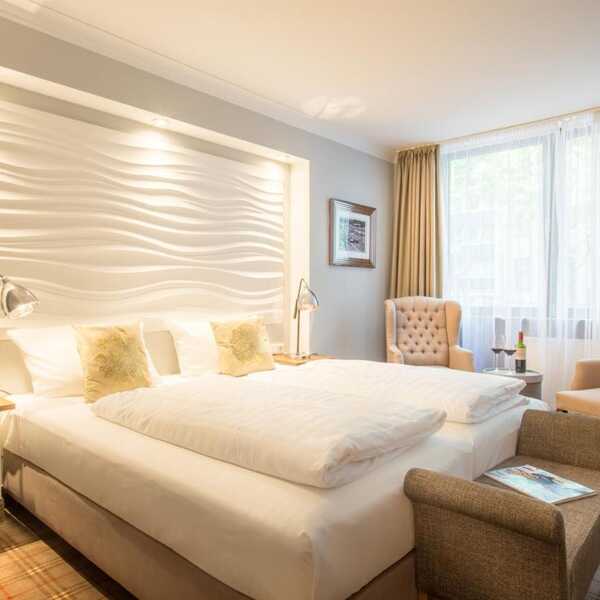 Ein helles, modernes Hotelzimmer mit Doppelbett, eleganten Sesseln und einem großen Fenster, das viel Licht hereinlässt.
