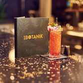 A vibrant cocktail garnished with flowers sits beside a menu on a sleek bar countertop at Bar Botanik.