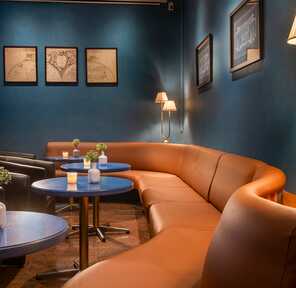 A cozy bar features a curved leather sofa, blue tables with candles, and wall art creating an inviting atmosphere.