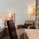 A well-lit superior room features a cozy armchair, elegant lighting, and a neatly made bed beside striped curtains.