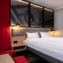 A modern hotel room features a cozy double bed, stylish red accents, and warm lighting, creating a welcoming atmosphere.