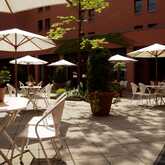 Sunny terrace with white tables and chairs under large umbrellas, surrounded by lush potted plants and red brick walls.