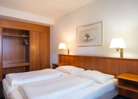 A cozy apartment bedroom features a neatly made bed, wooden closet, and soft lighting from bedside lamps.