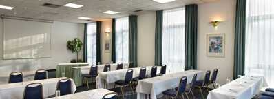 A well-lit meeting room features rows of tables and chairs set up for a conference with a projection screen at the front.