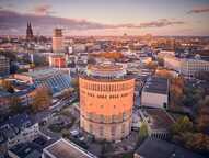 Der historische Wasserturm in Köln wird von oben mit der Skyline der Stadt im Hintergrund bei Sonnenuntergang gezeigt.