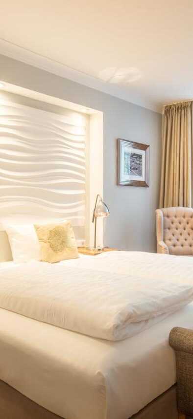 Ein helles, modernes Hotelzimmer mit Doppelbett, eleganten Sesseln und einem großen Fenster, das viel Licht hereinlässt.