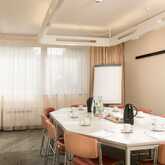 A fully equipped conference room at the Best Western Hotel Achim Bremen with a large, bright, hexagonal conference table and matching orange-coloured chairs. On the table are cups, water bottles, coffee pots, notepads and pens. A flipchart and a window fr