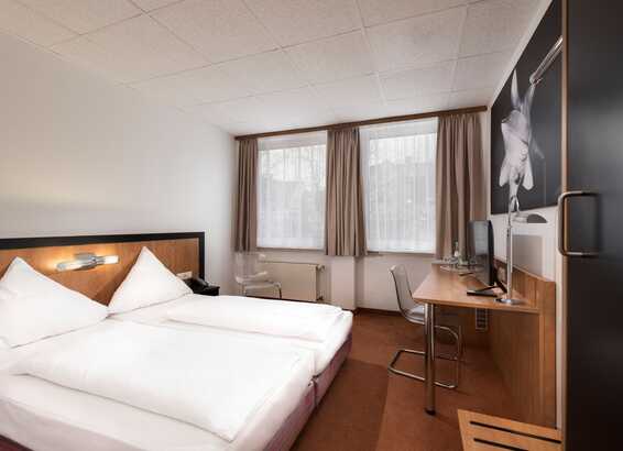 A modern double room features a neatly made bed, a desk with a computer, and two large windows with beige curtains.