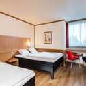 A cozy bedroom features two neatly made beds, a red chair by the window, and warm wooden accents creating a welcoming atmosphere.