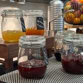 Jars of various fruit jams are neatly arranged on a breakfast table, alongside a basket of fresh apples and oranges.