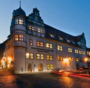 The illuminated exterior of Hotel Quedlinburger Stadtschloss glows warmly at dusk with blurred car lights on the cobblestone street.