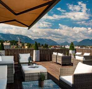 A rooftop terrace with wicker furniture offers a stunning mountain view under a partly cloudy sky.