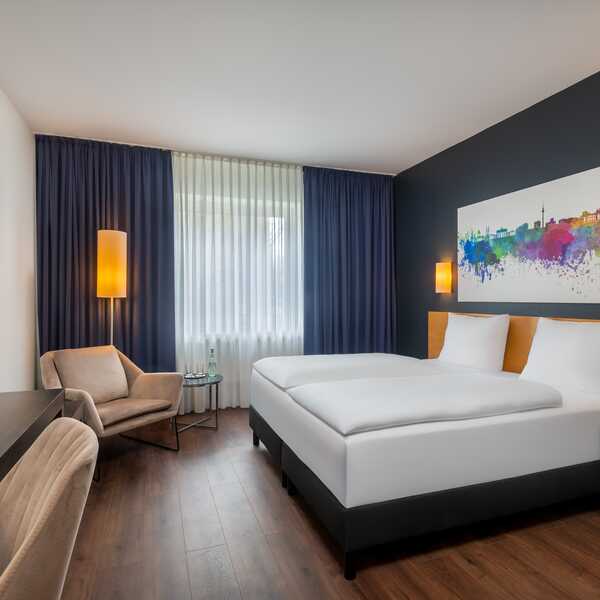 Ein modernes Hotelzimmer mit einem Doppelbett, farbenfroher Wandkunst und einem gemütlichen Sessel vor dunklen Vorhängen.