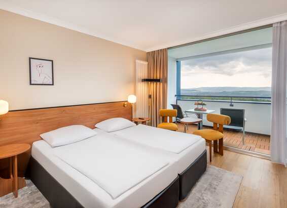 A modern hotel room with twin beds and a balcony overlooking a scenic landscape, featuring stylish decor and warm lighting.
