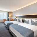 A modern hotel room features two neatly made beds with blue accents, a desk, and a window letting in natural light.