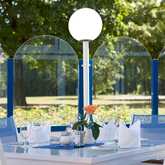 A beautifully set outdoor terrace table features elegant glassware and a vibrant flower centerpiece against a lush garden backdrop.